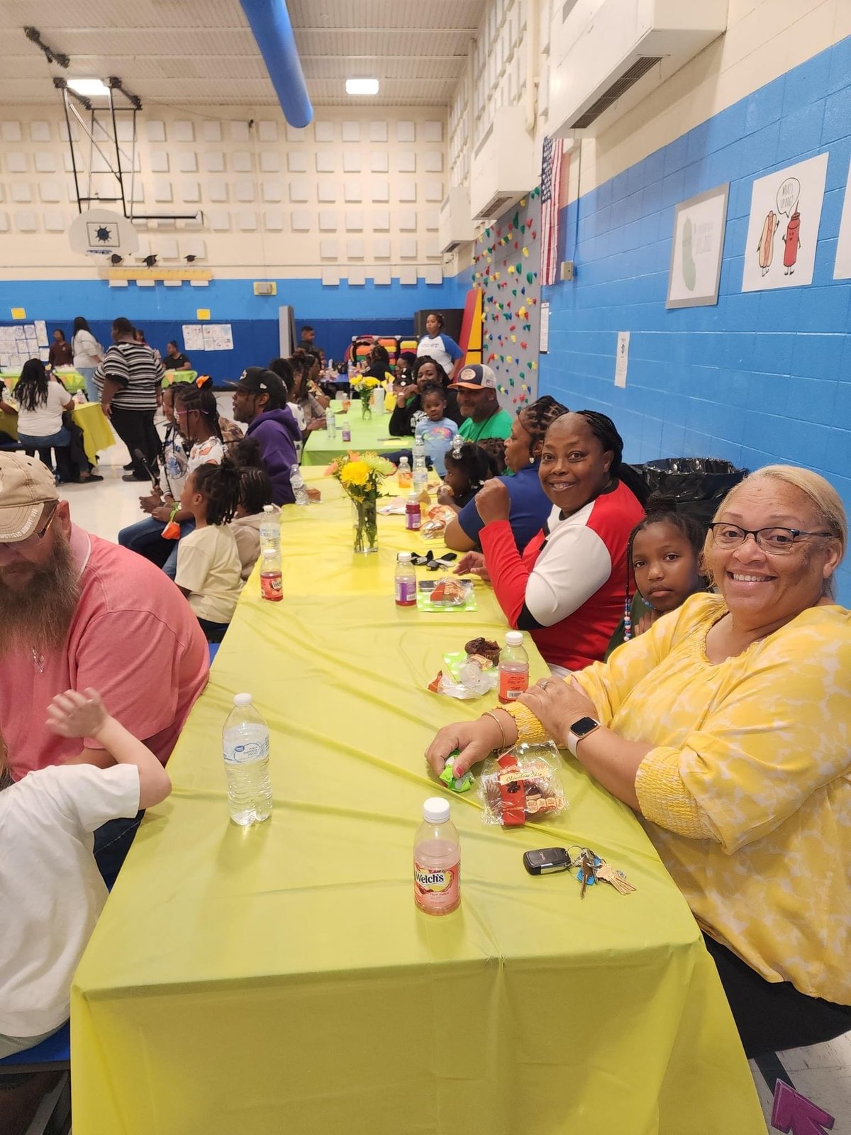 Children and adults sitting at long yellow tables eating and socializing in a school cafeteria with blue walls