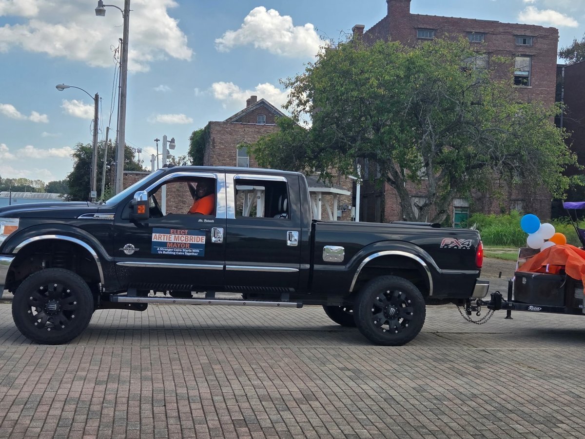 Black pickup truck with Elect Artie McBride Mayor sign parked on brick pavement with city buildings in the background
