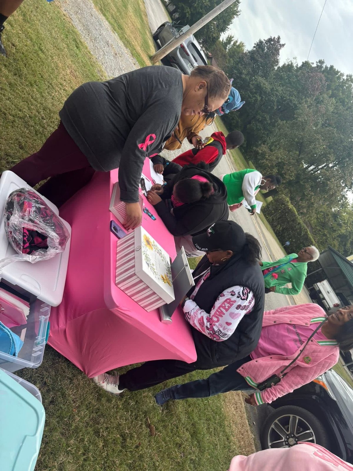 Adult and children at a pink vendor table at an outdoor community event in a residential yard