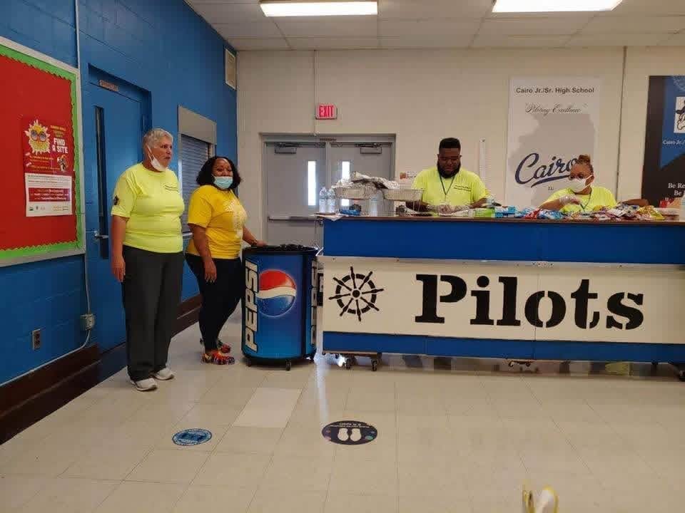 School cafeteria with staff in yellow shirts behind a counter labeled Pilots, serving food to students