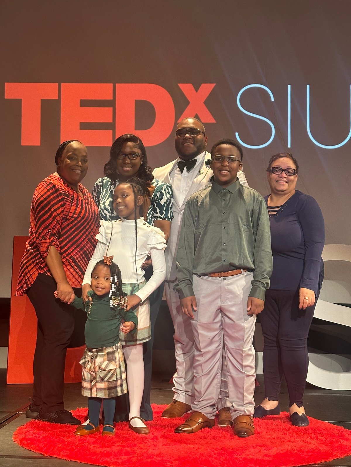 Group of six people standing on the red carpet at TEDxSIU, smiling together with the red TEDx letters behind them