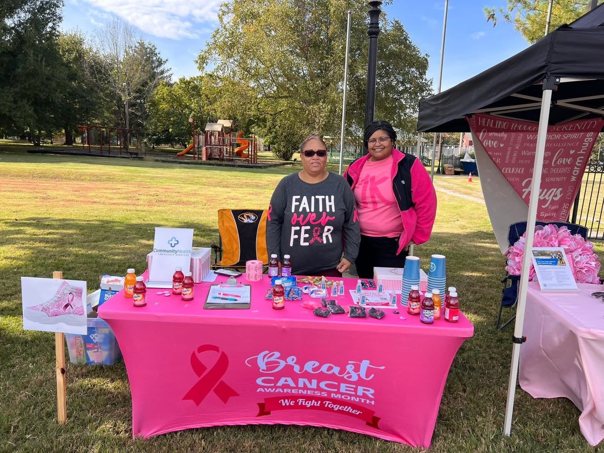 Two women at a breast cancer awareness fundraiser booth with pink table, products, and signage in a park setting