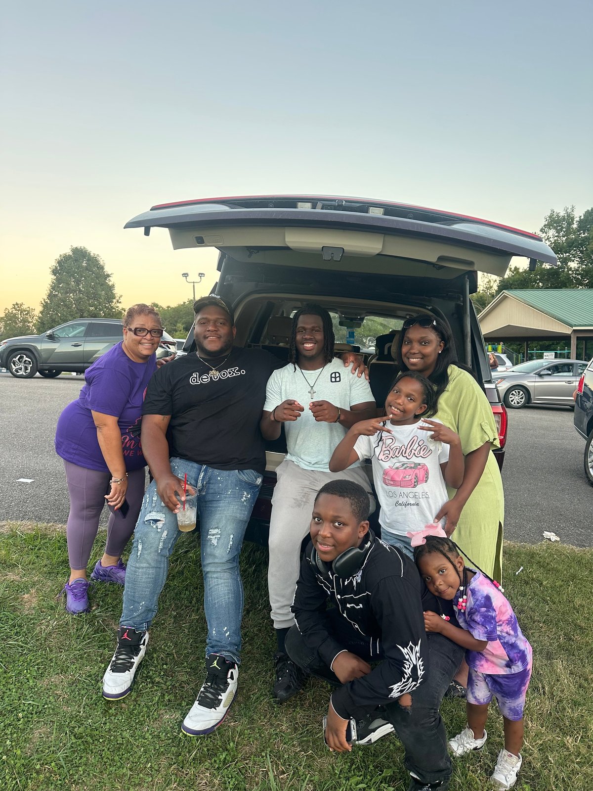 Group of seven people of varying ages smiling at the open trunk of an SUV in a parking lot during daytime
