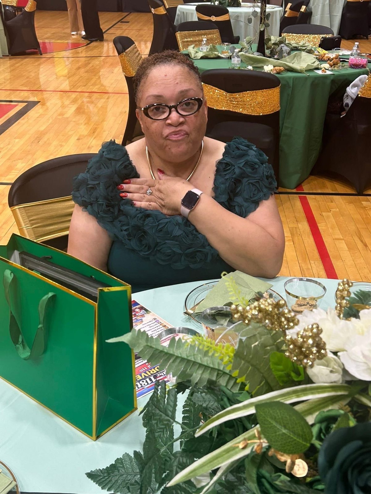 Woman with glasses wearing a green ruffled top sits at a table with jewelry displays at an event