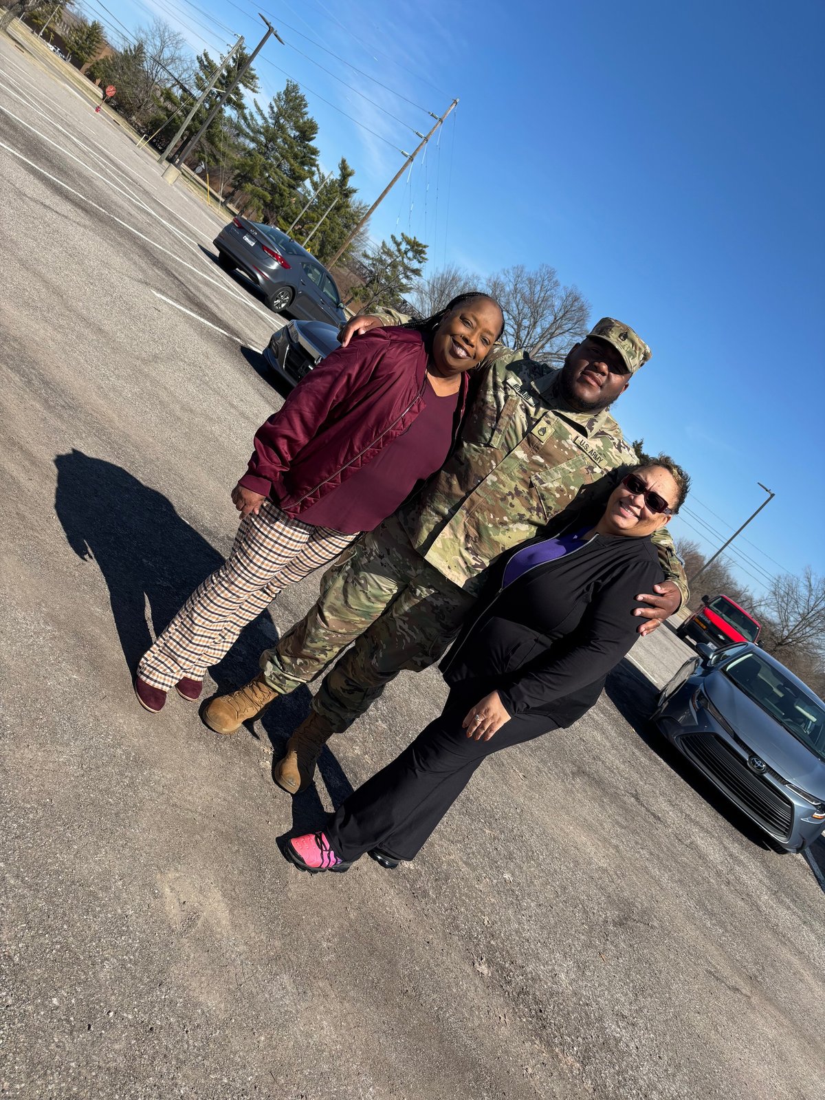 Three people posing together on a paved road with vehicles and bare trees in the background