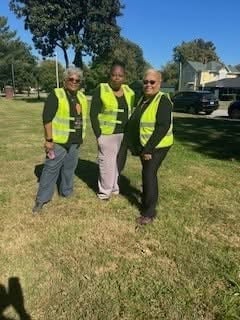 Three workers in yellow safety vests stand on a grass field with trees and vehicles in the background