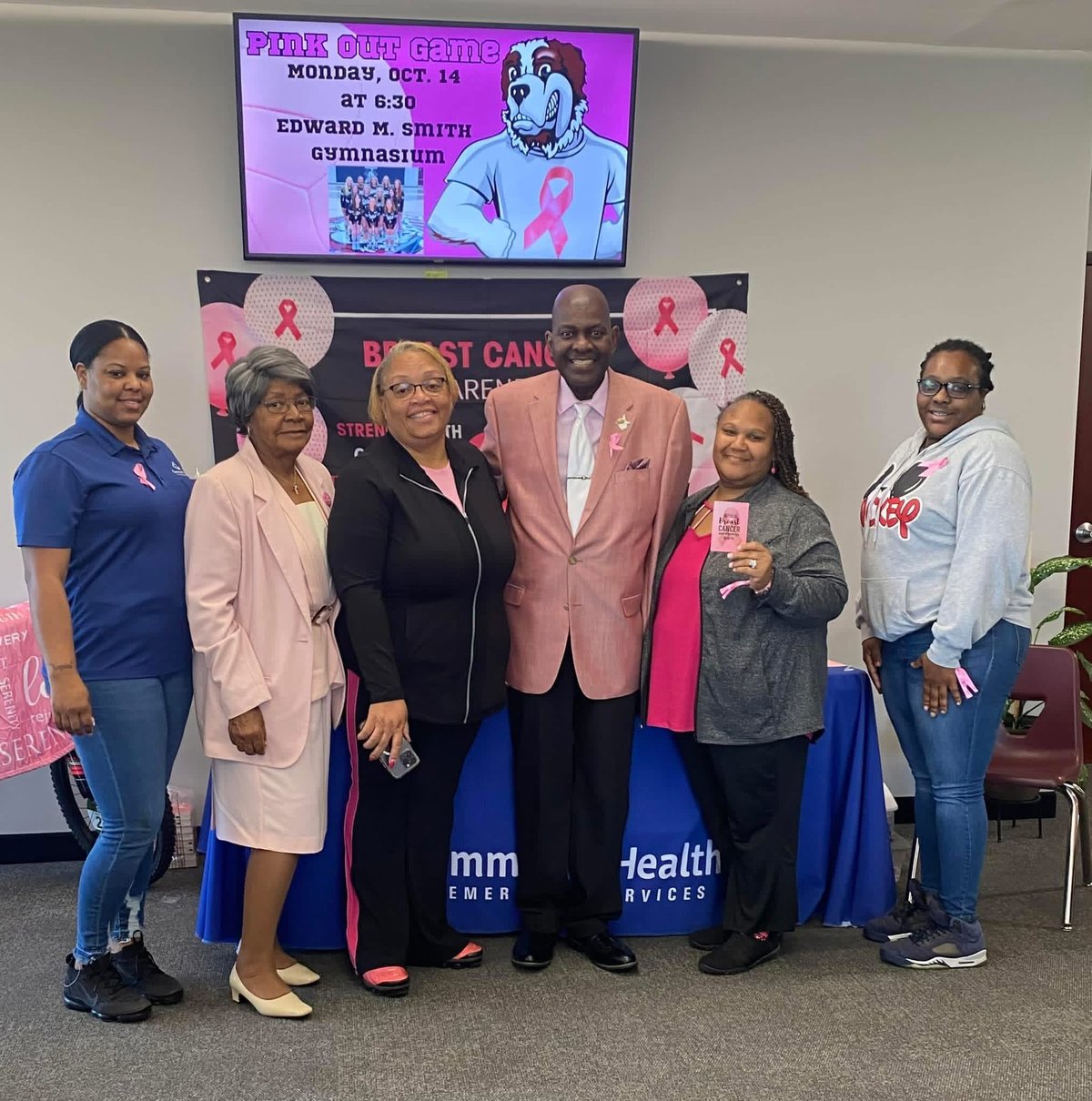 Group of six people posing in front of a pink ribbon breast cancer awareness banner and event screen