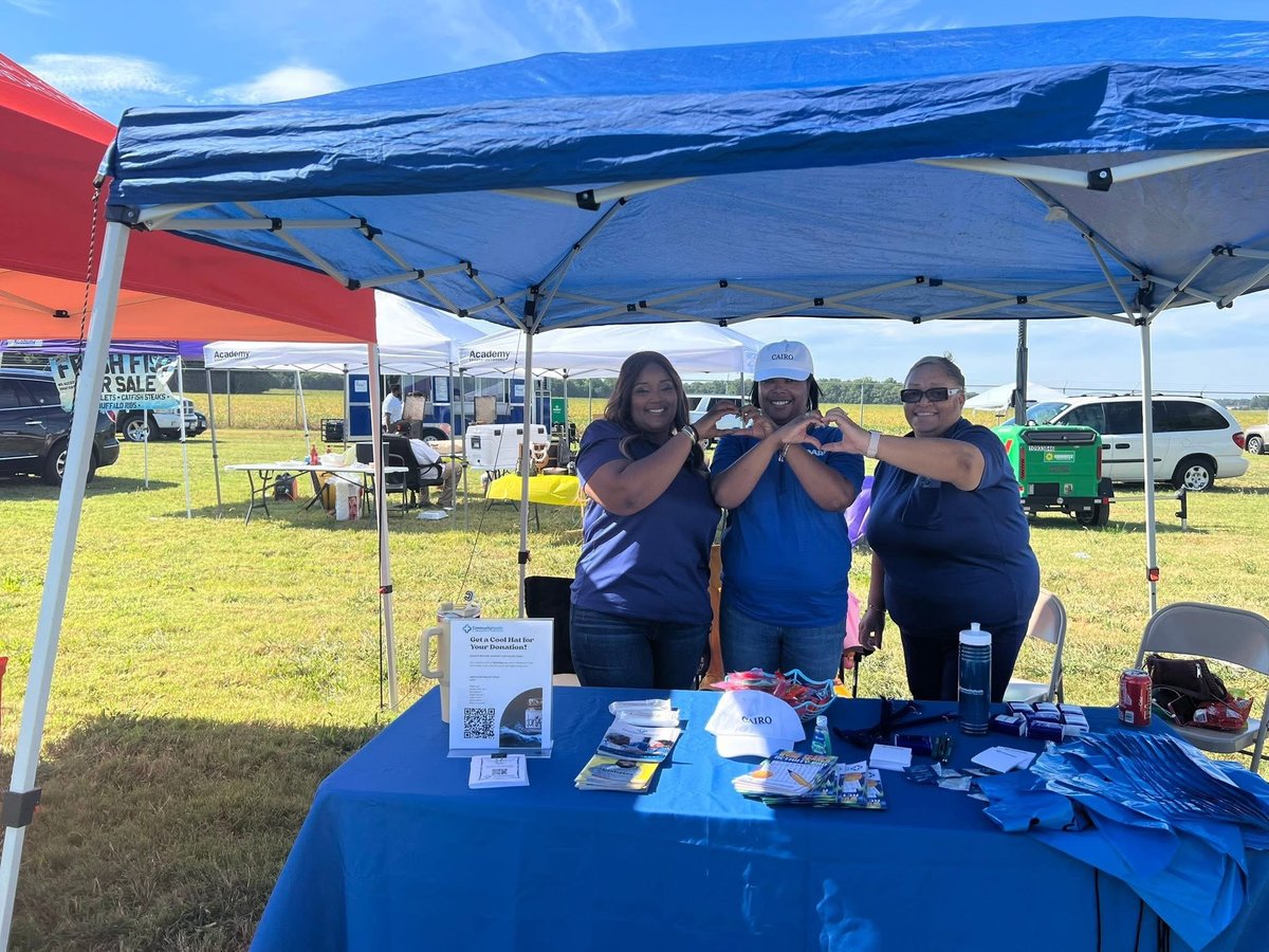 Three women in blue shirts standing behind a blue table under a blue pop-up canopy at an outdoor event in a field