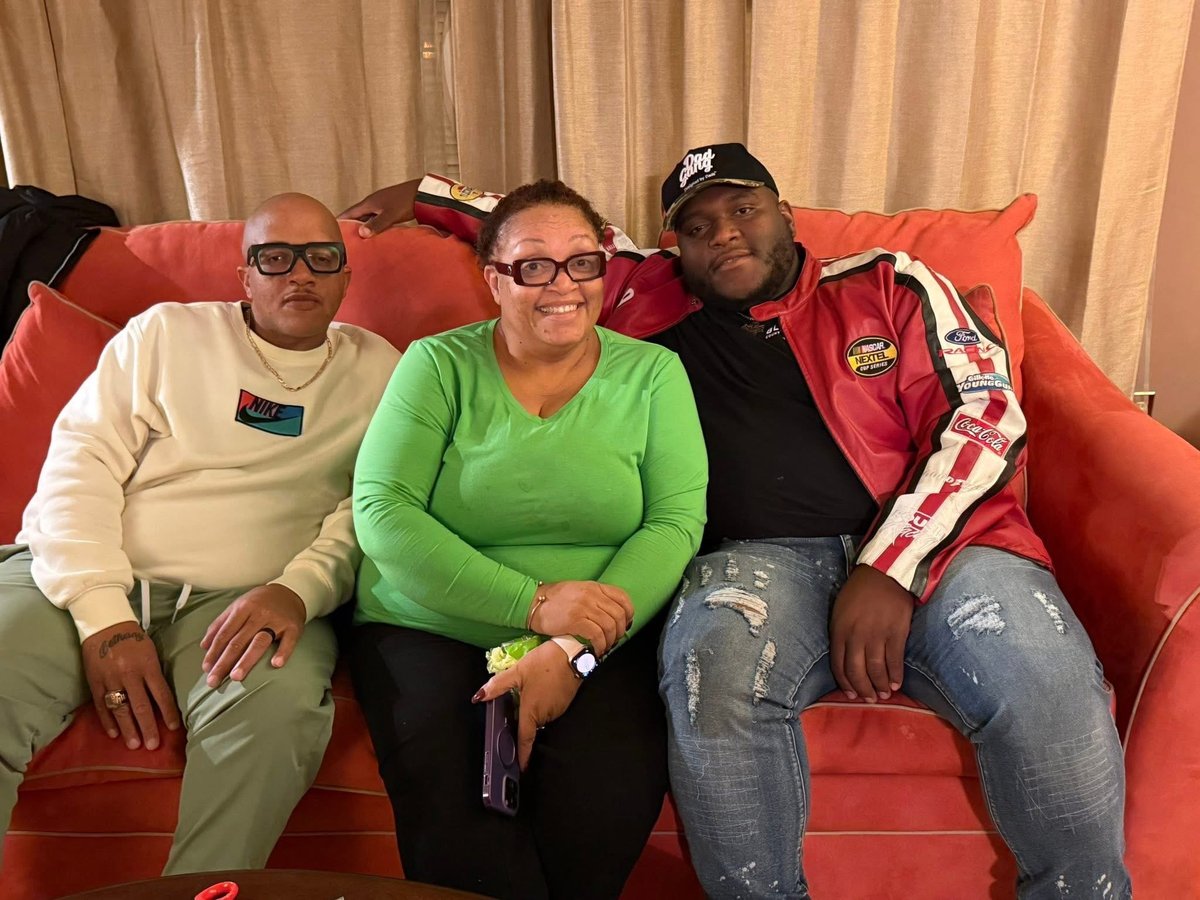 Artie in a green top sits on a red couch between her two sons, all three smiling at the camera in a casual indoor setting