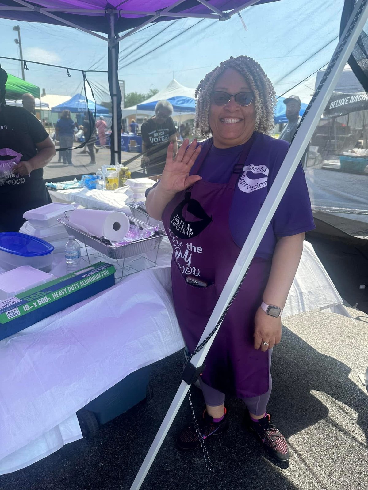 Woman wearing purple shirt waving at camera inside a vendor tent at an outdoor event