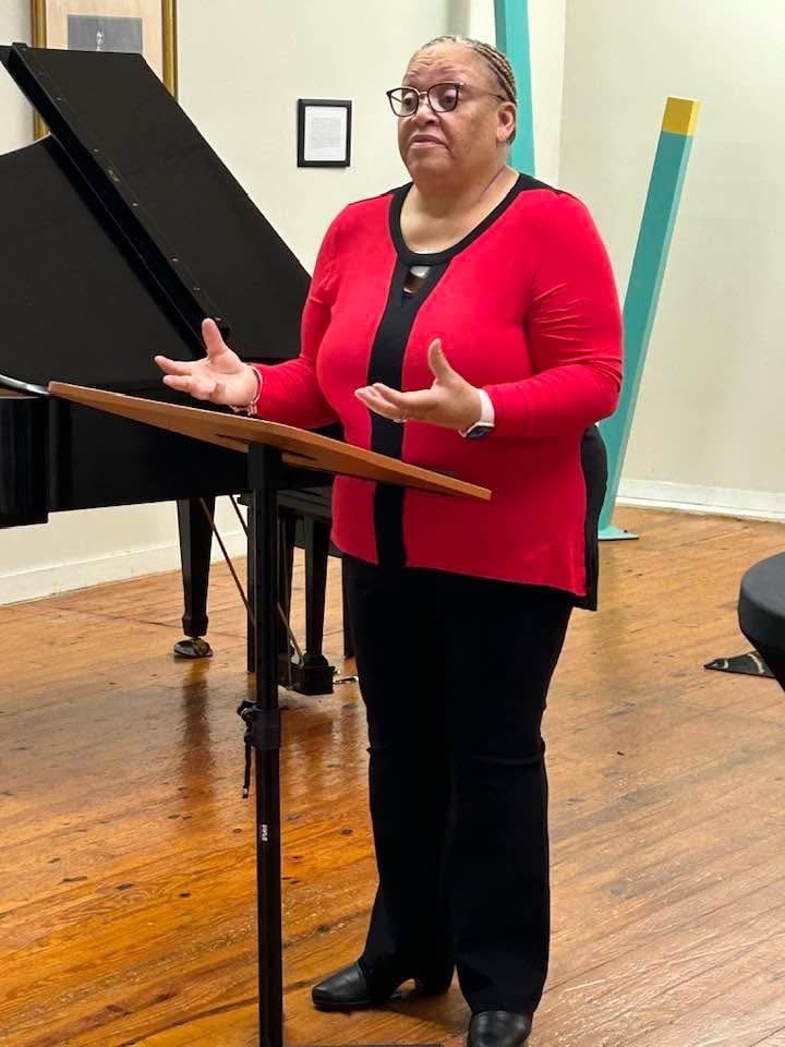 Woman in red cardigan speaking at a podium next to a grand piano in a music studio