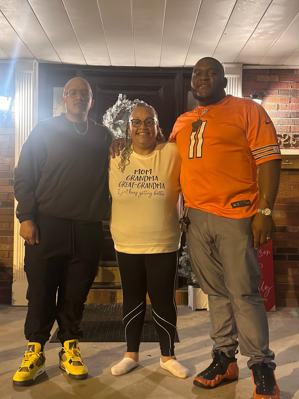 Three people standing together indoors, with the woman in the center wearing a yellow shirt that reads 'Mom Grandma Great-Grandma I just keep getting better'