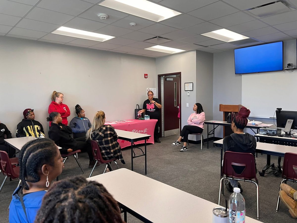 Indoor classroom or community room with diverse group of people seated in chairs, with instructors at a pink table and blue wall-mounted screen