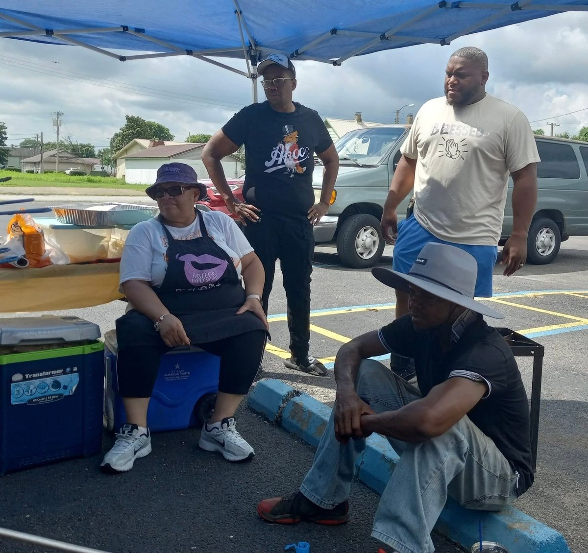 Four men gathered under a blue canopy in a parking lot, with coolers and vehicles visible in the background